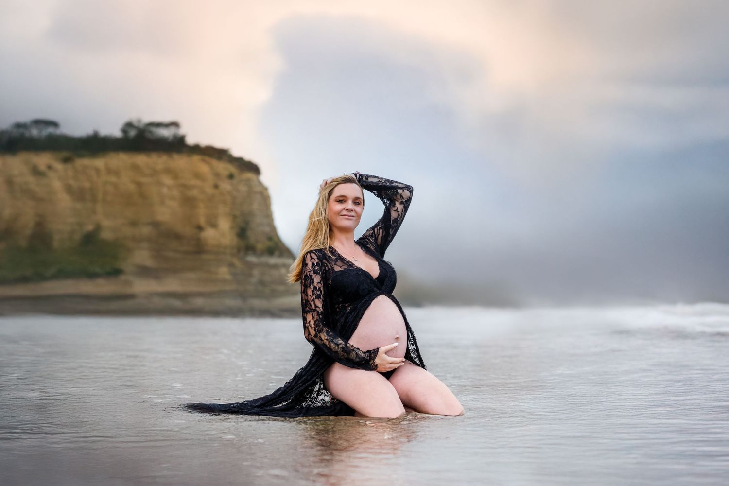 Pregnant woman kneeling in ocean water wearing a flowing black robe during a beach maternity session by an Auckland photographer