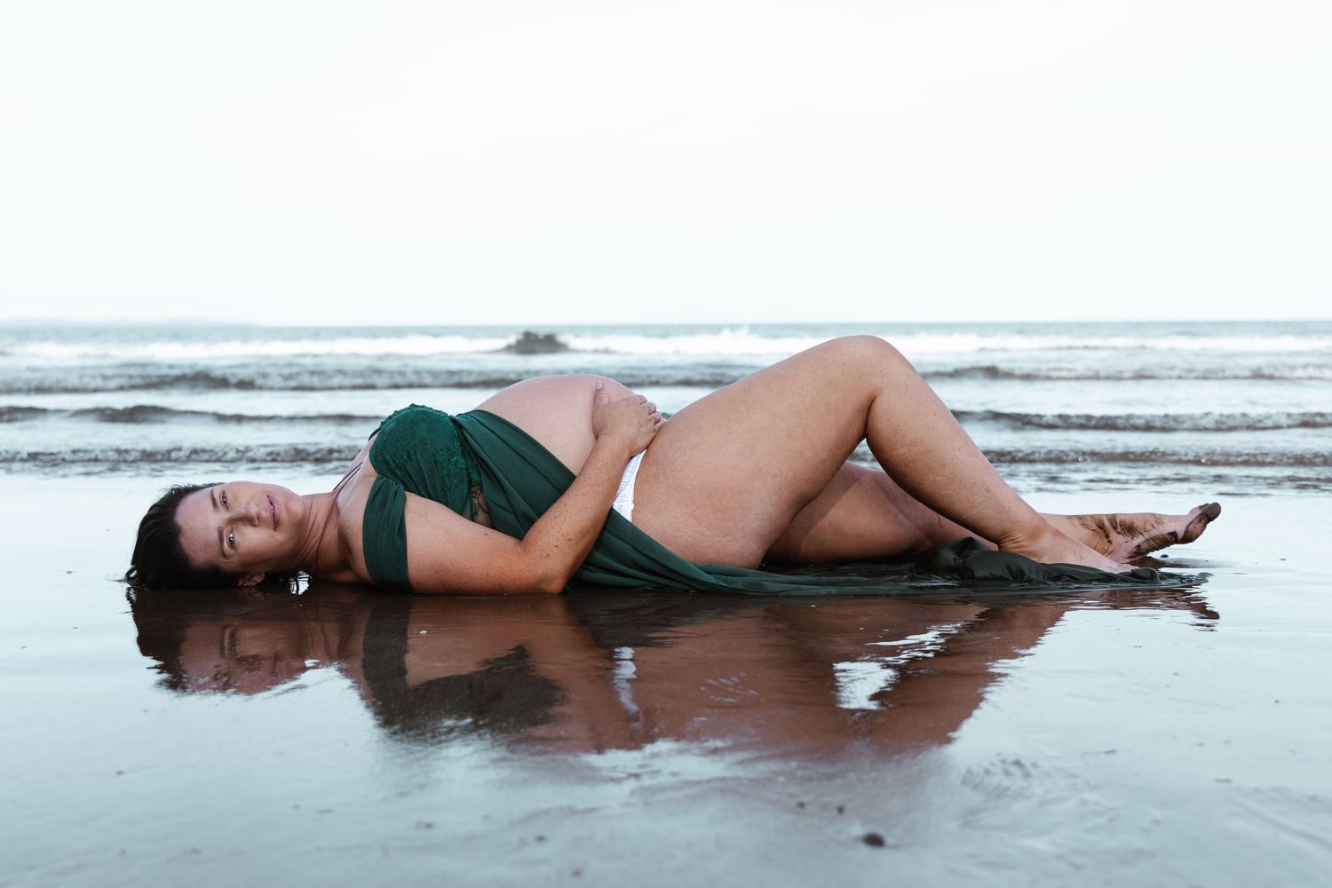 Pregnant woman lying on wet sand at an Auckland beach in a green outfit, photographed by an Auckland photographer.