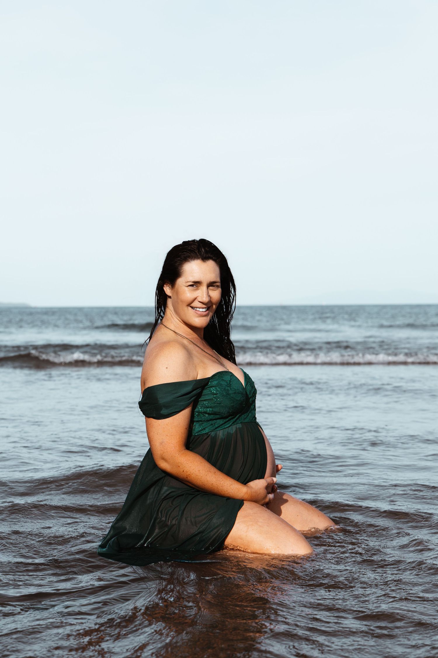 Expectant mother in a green dress seated in the sea at an Auckland beach, captured by an Auckland photographer.