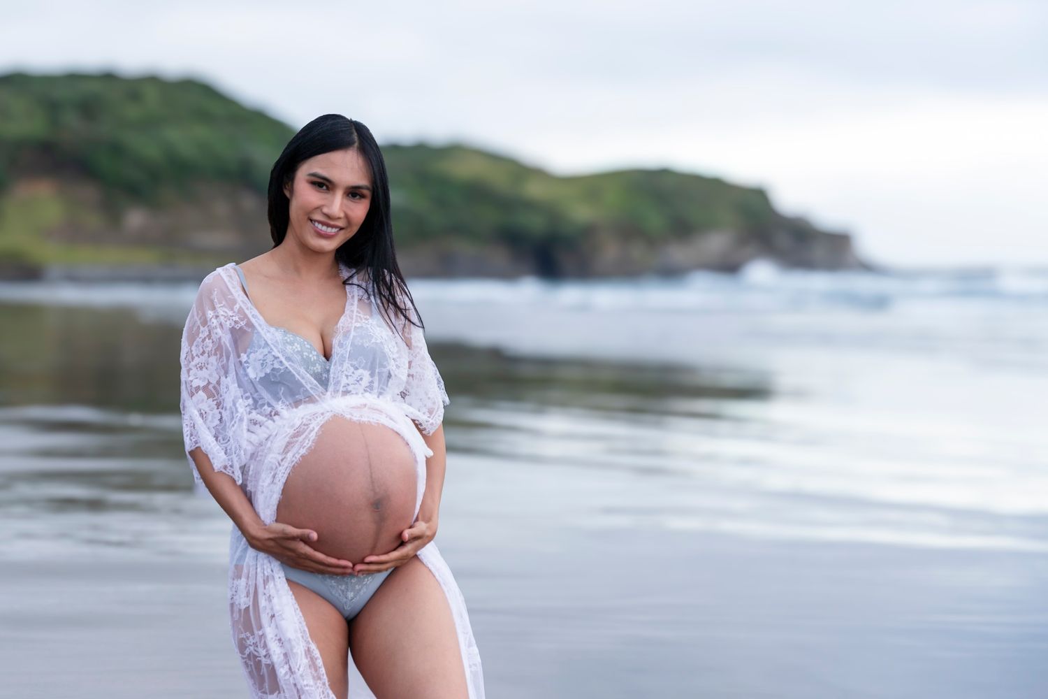 Muriwai Beach Maternity Photography: Elegant pregnant woman in white lace dress posing at the water's edge, Auckland West Coast.