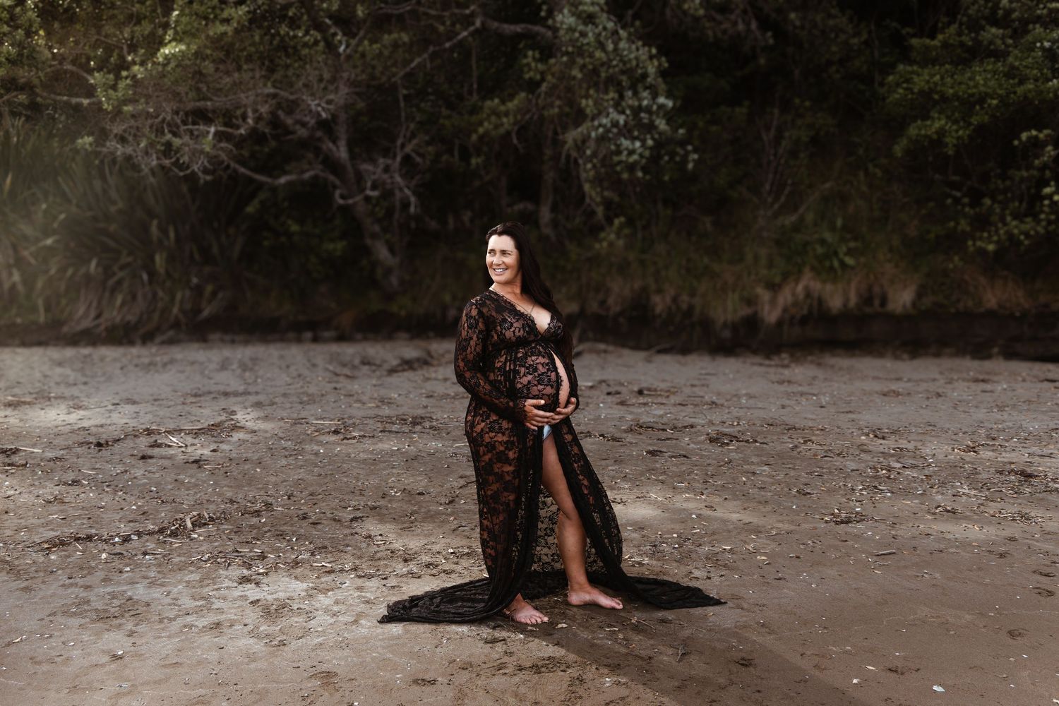 Pregnant woman standing barefoot on a sandy Auckland beach in a black flowing dress, photographed by an Auckland photographer.