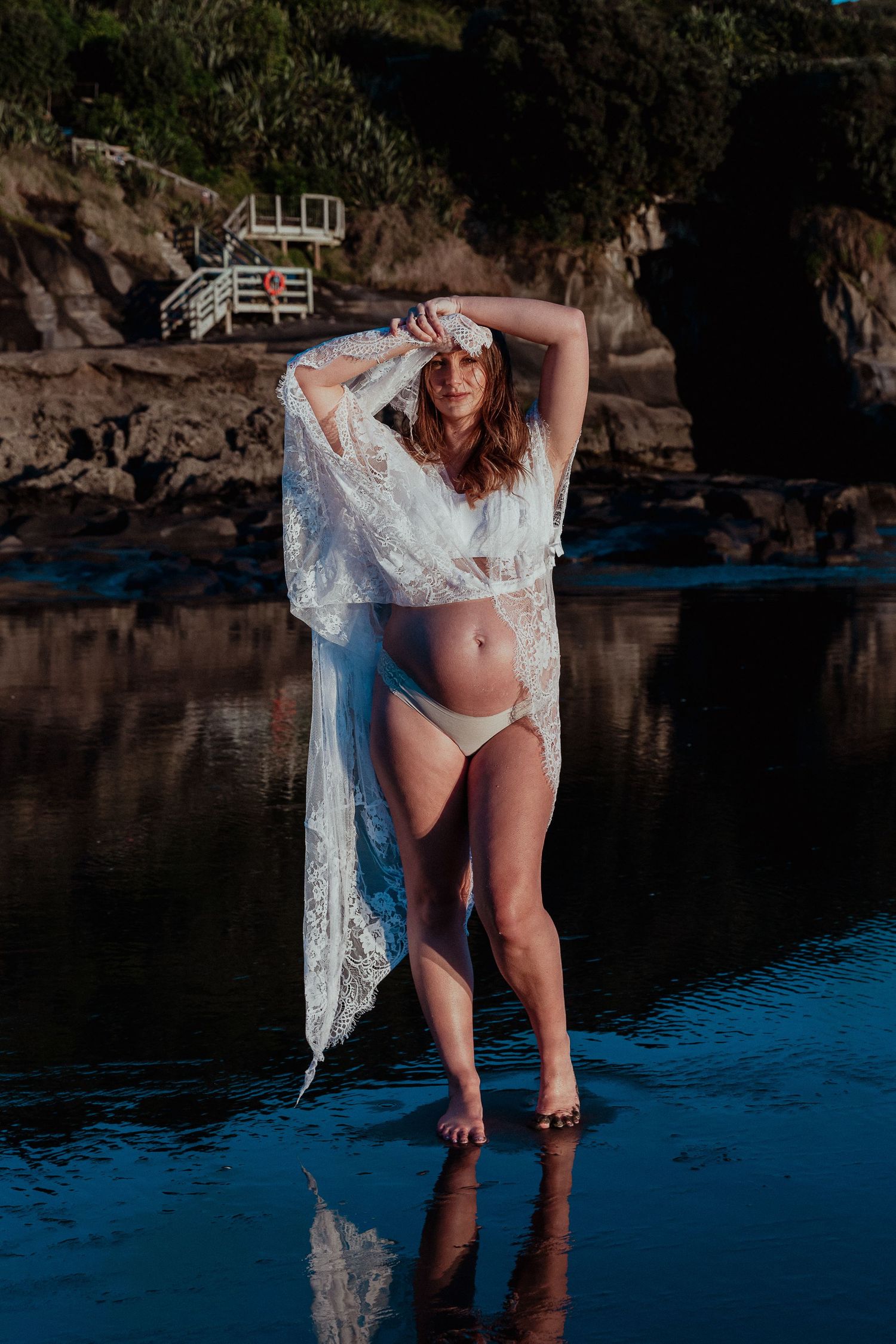 Muriwai Beach Maternity Session: Woman in dark dress posing against dramatic rocks at sunset, Auckland.