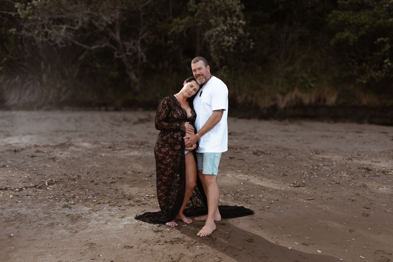 Pregnant woman and partner embracing on an Auckland beach, captured during a maternity shoot by an Auckland photographer.
