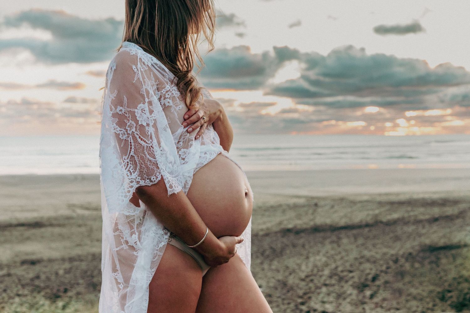 Muriwai Beach Maternity Photos: Pregnant woman in white lace dress standing on the beach, Auckland North Shore.