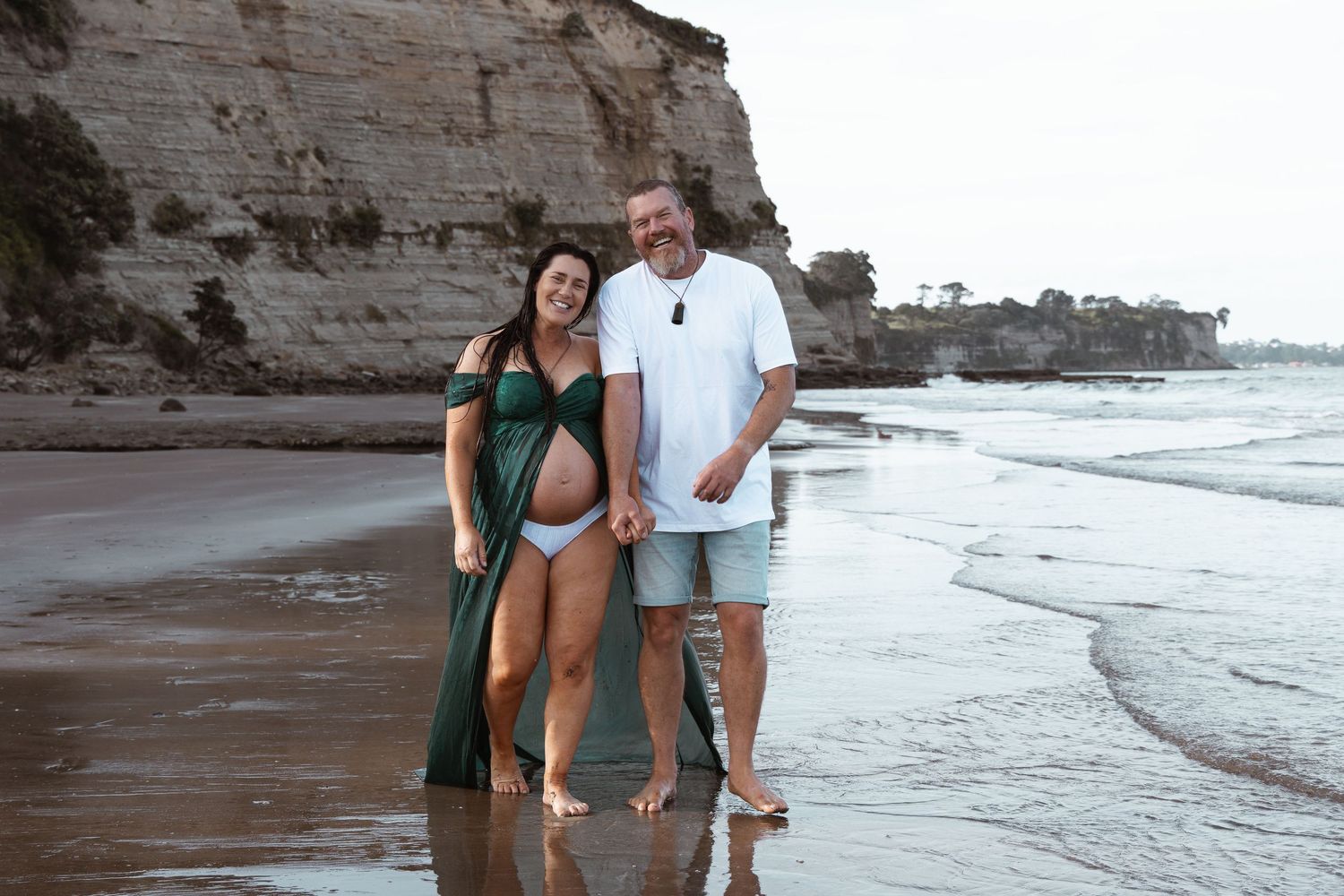 Expectant couple standing and laughing together on an Auckland beach during a maternity shoot by an Auckland photographer.