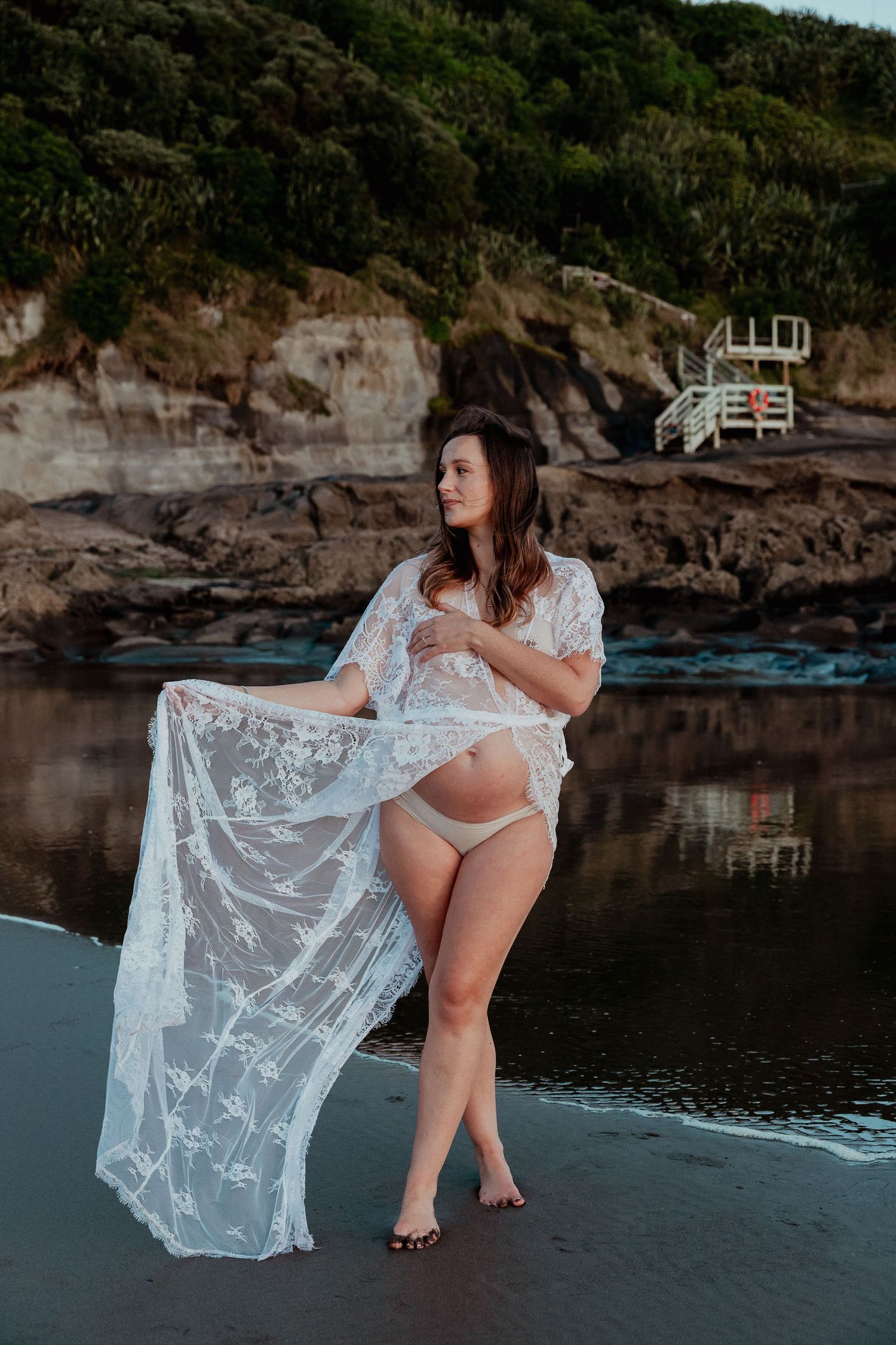 Auckland Maternity Photographer: Elegant pregnant woman in white lace dress at Muriwai Beach sunset.