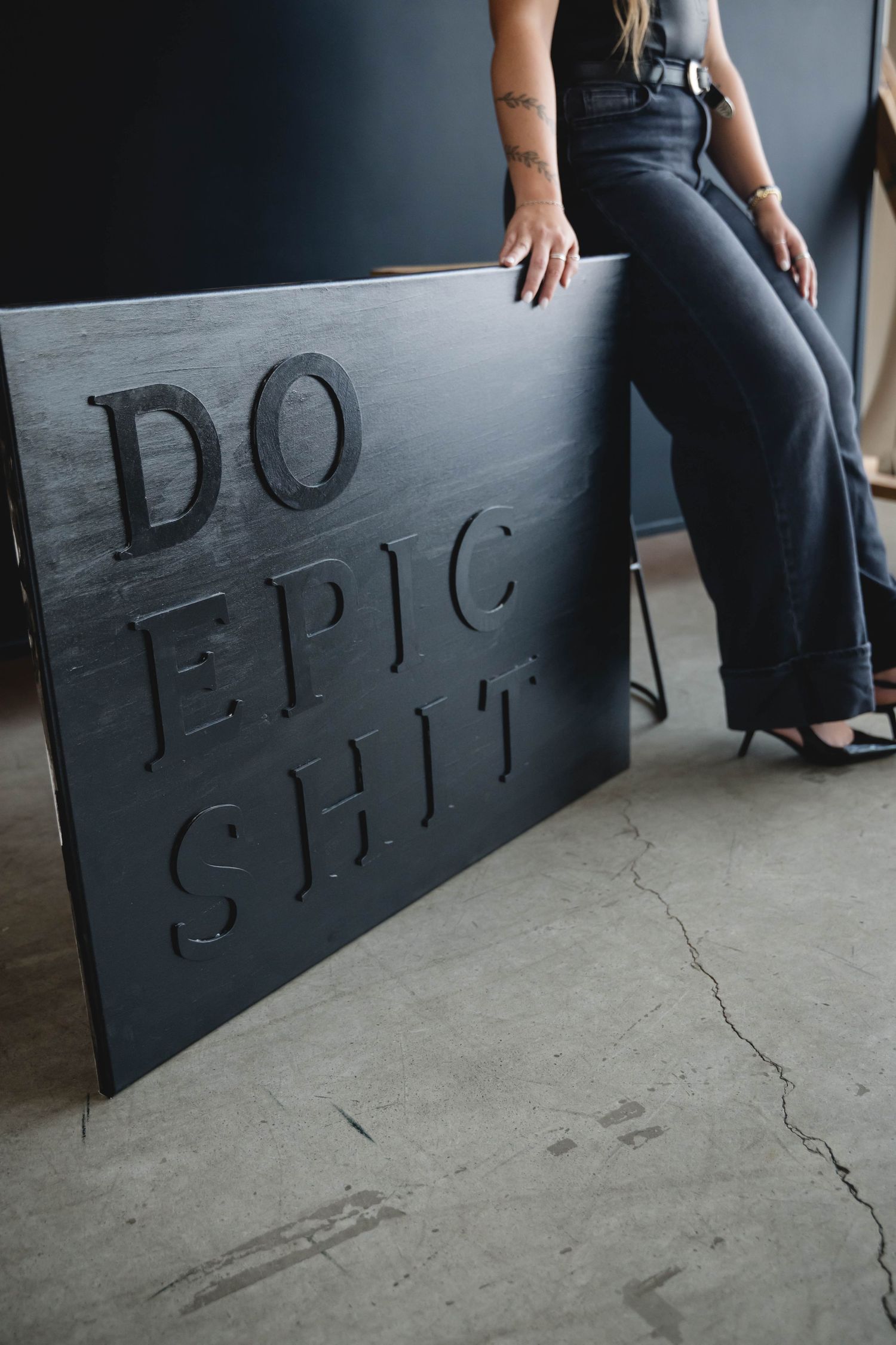 Person wearing black jeans sits on floor next to dark wall with text written in chalk.