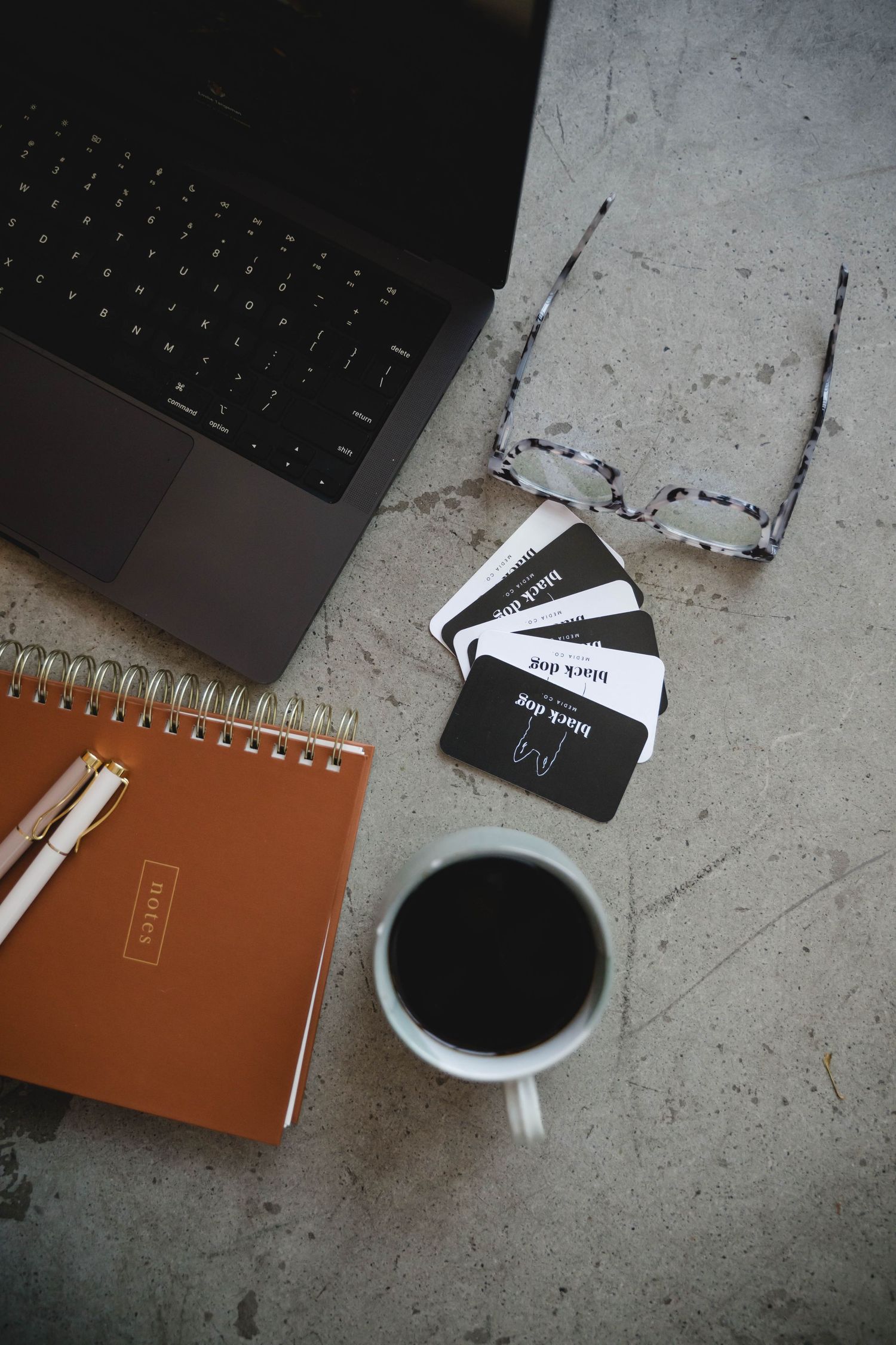 Flatlay workspace with laptop, brown notebook, glasses, black coffee, and business cards on concrete surface.