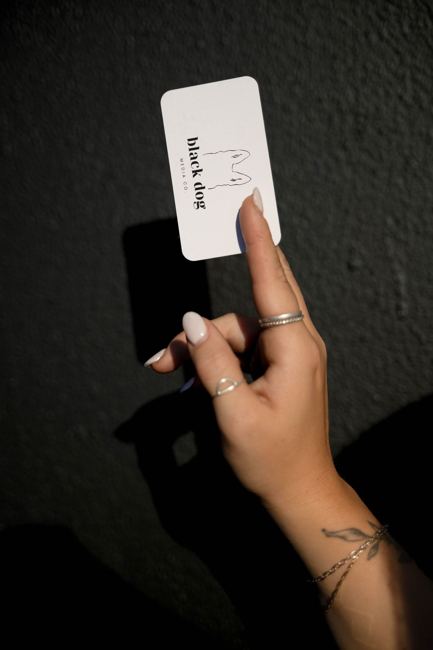 A hand with a small tattoo holds up a Black Dog Media business card against a dark background.