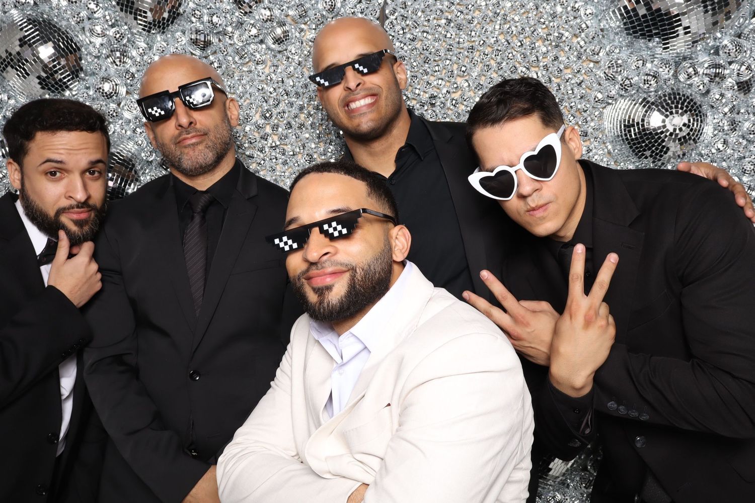 Five people in formal attire and sunglasses pose against sparkly silver backdrop.
