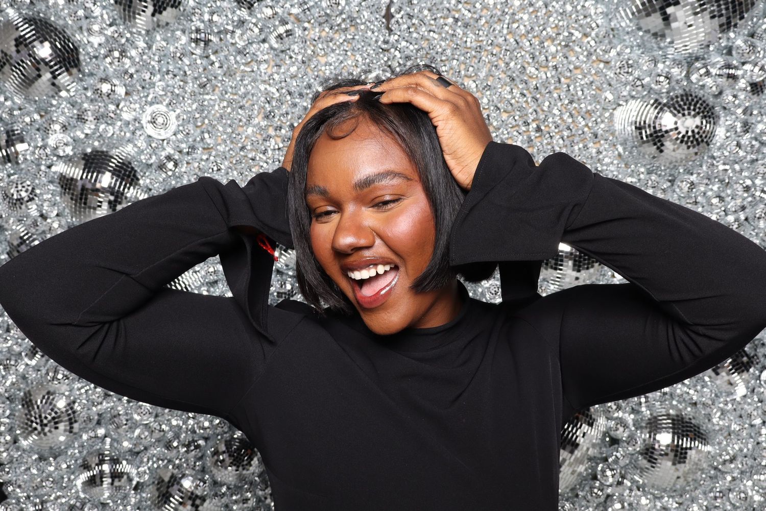 Person in black sweater laughs joyfully against sparkly silver background.