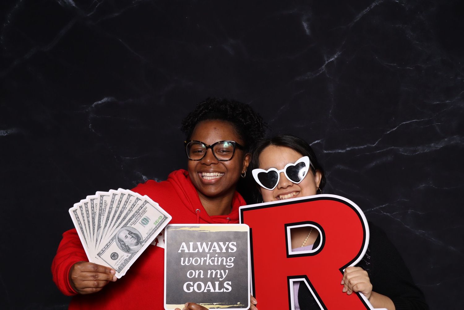 Two people pose with cash and motivational sign in front of black backdrop at photo booth.