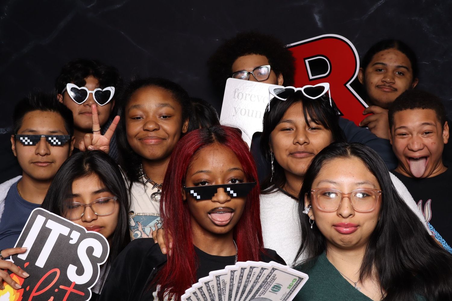 Diverse group showing off cash and props while posing together in photo booth with letter R sign.