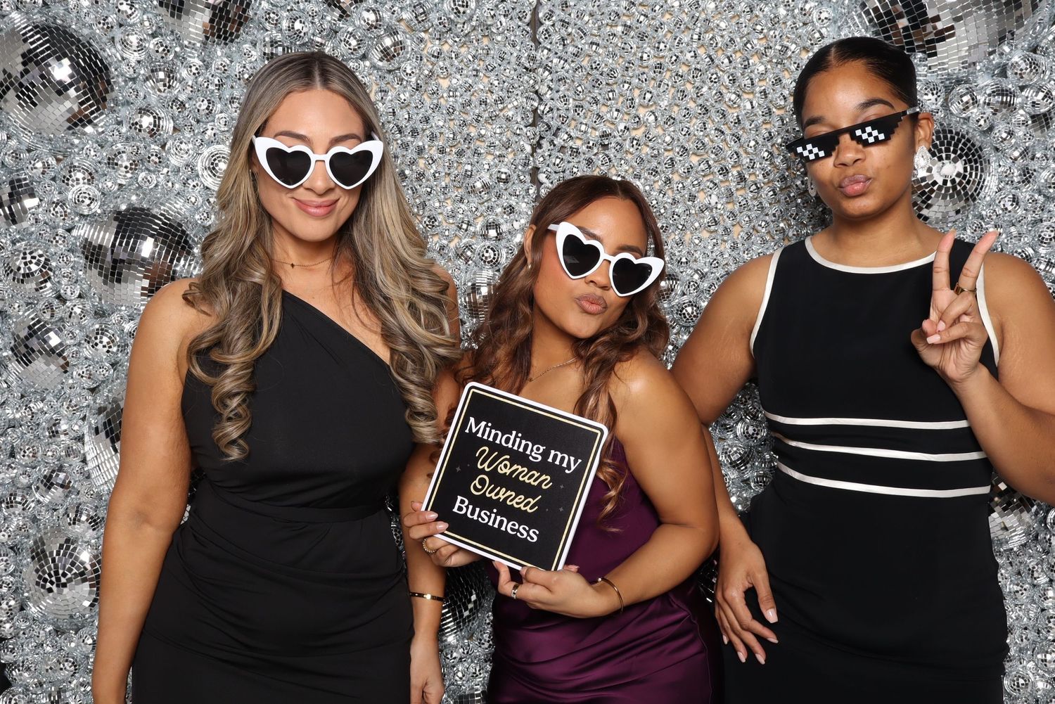 Three friends pose with sunglasses and signs against sparkly silver background.
