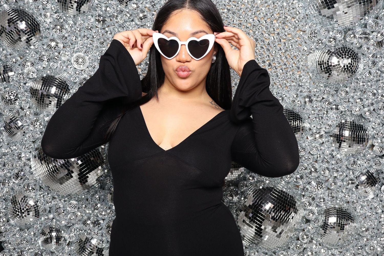 Person in black dress adjusts white sunglasses while posing against silver backdrop.
