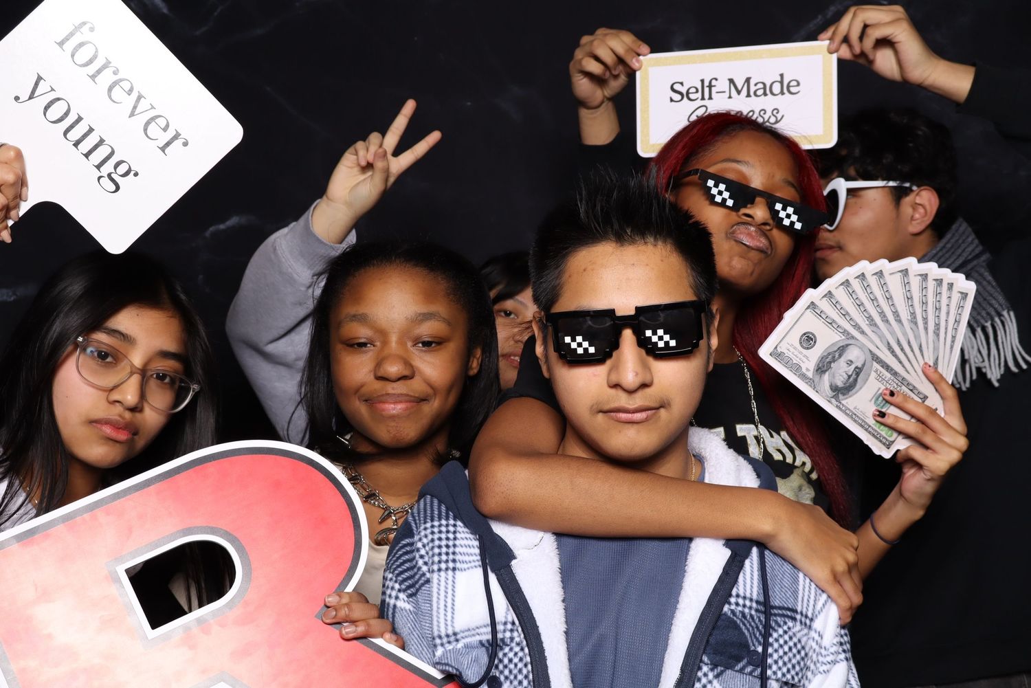 Group poses with sunglasses, cash and signs in a fun photo booth setting against black background.