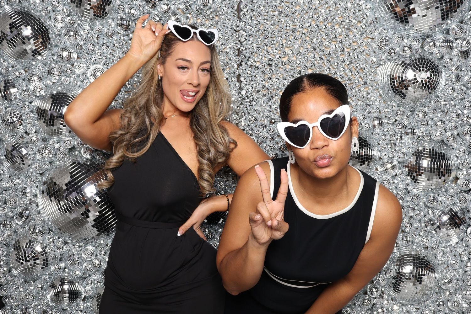 Two friends in black outfits pose with sunglasses against silver sequin background.