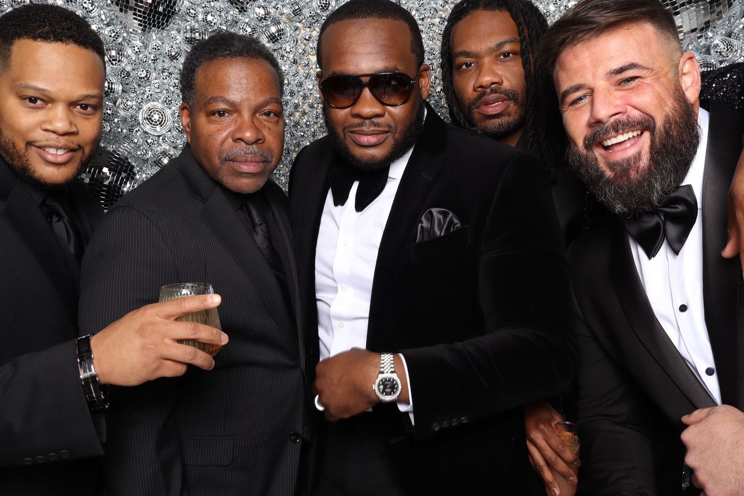 Group of men in formal black attire pose together against silver sequin backdrop.