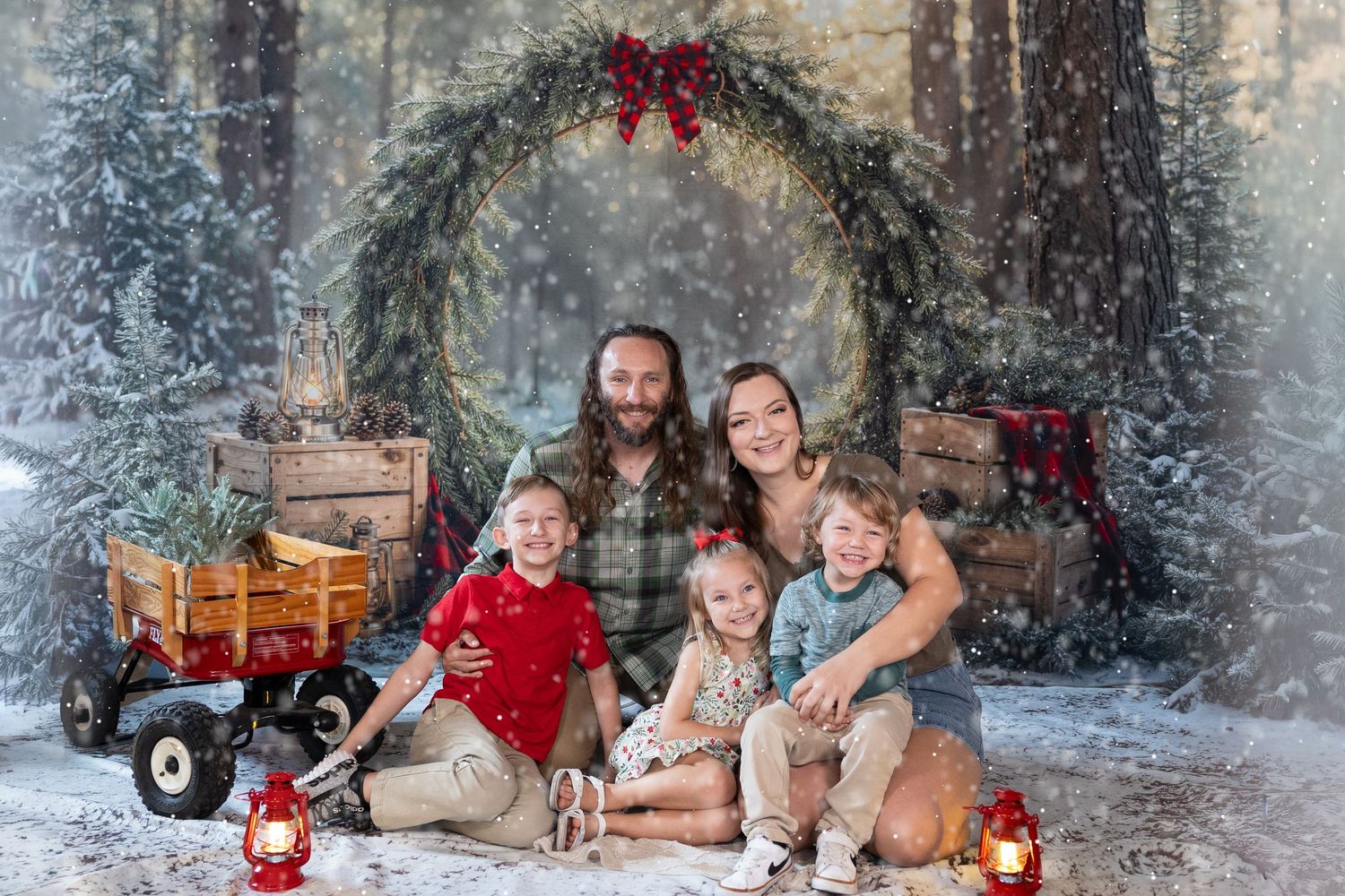 Family of five posing for a festive Christmas photo in a snowy forest setting with wreaths and holiday decorations.