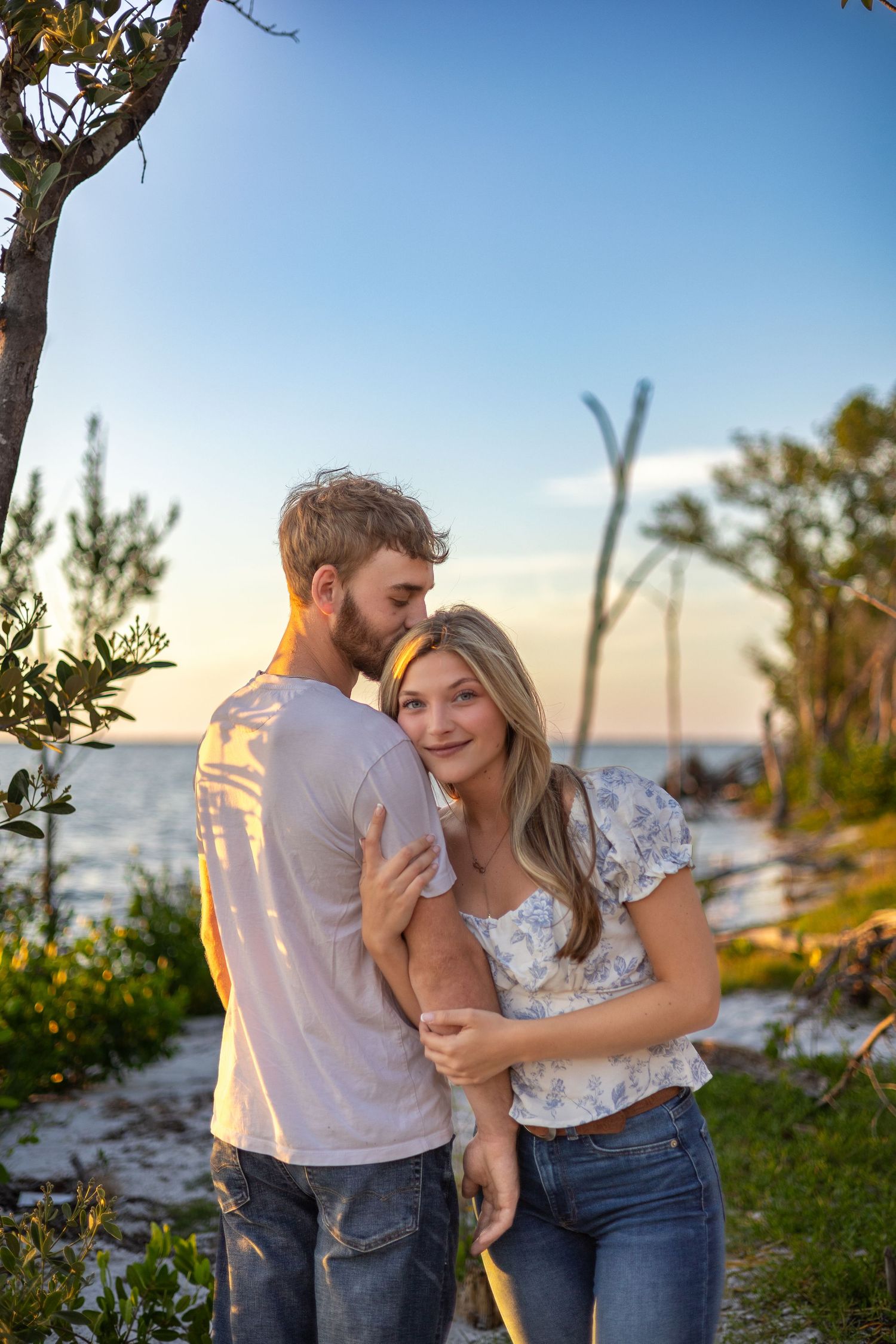 A couple embraces at sunset near a sandy beach with trees, the woman smiling warmly in a floral top and jeans.