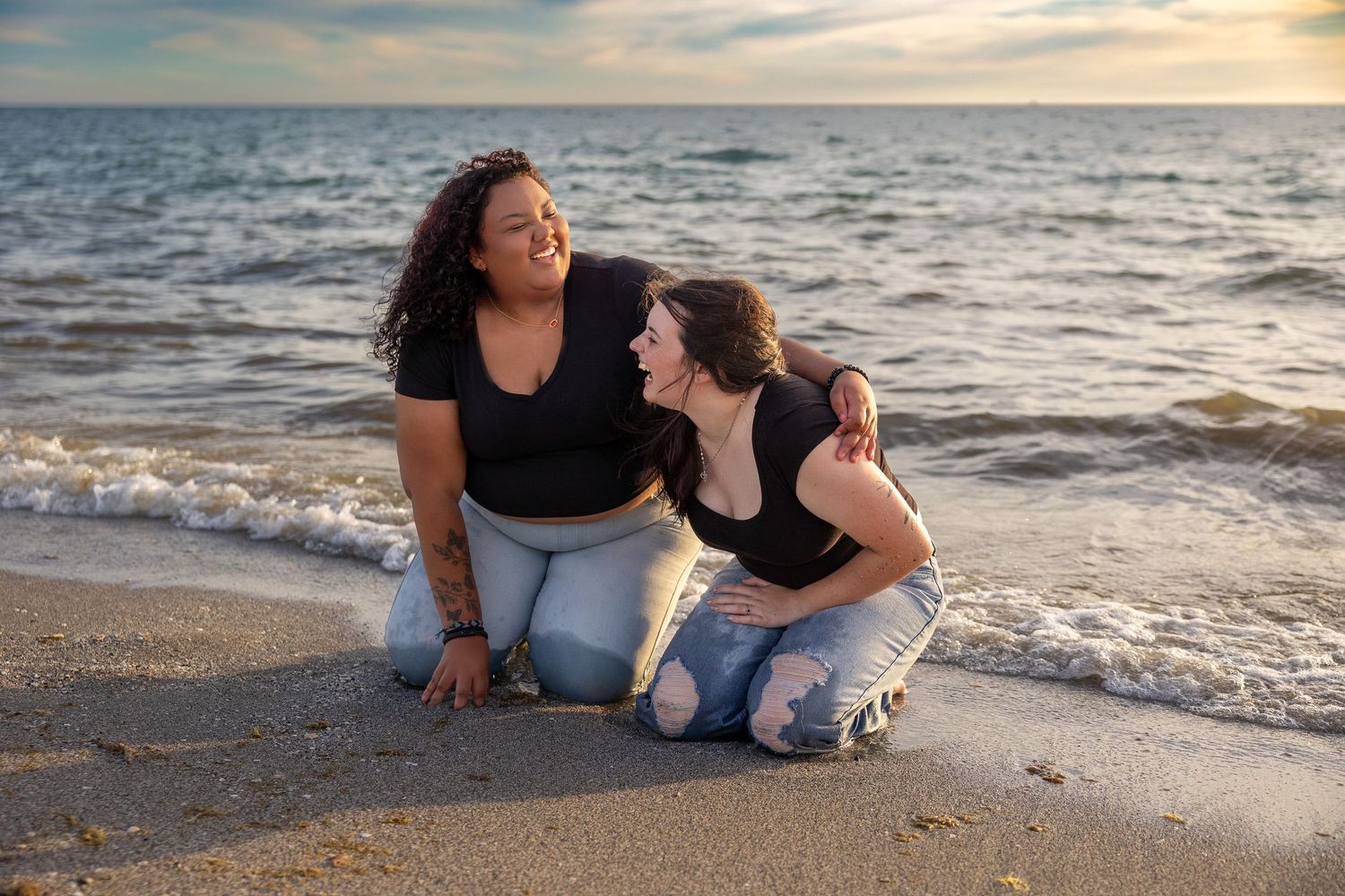 Two women laughing together while kneeling on wet sandy beach at sunset.