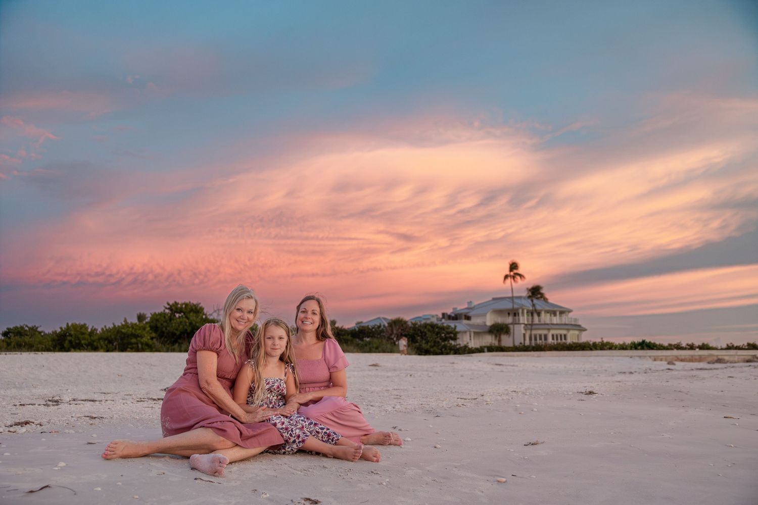 Three women in pink dresses pose on white sand beach at sunset with lighthouse backdrop.