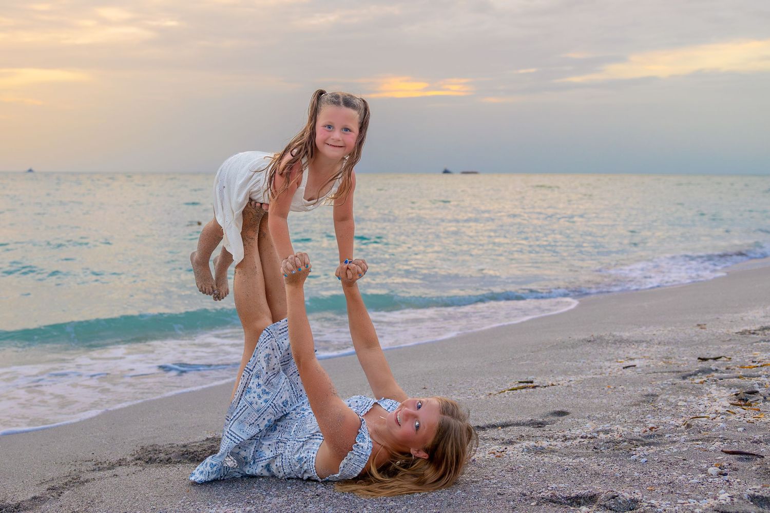 Mother lying on beach lifting young daughter in air at sunset, both smiling in white dresses by turquoise ocean.