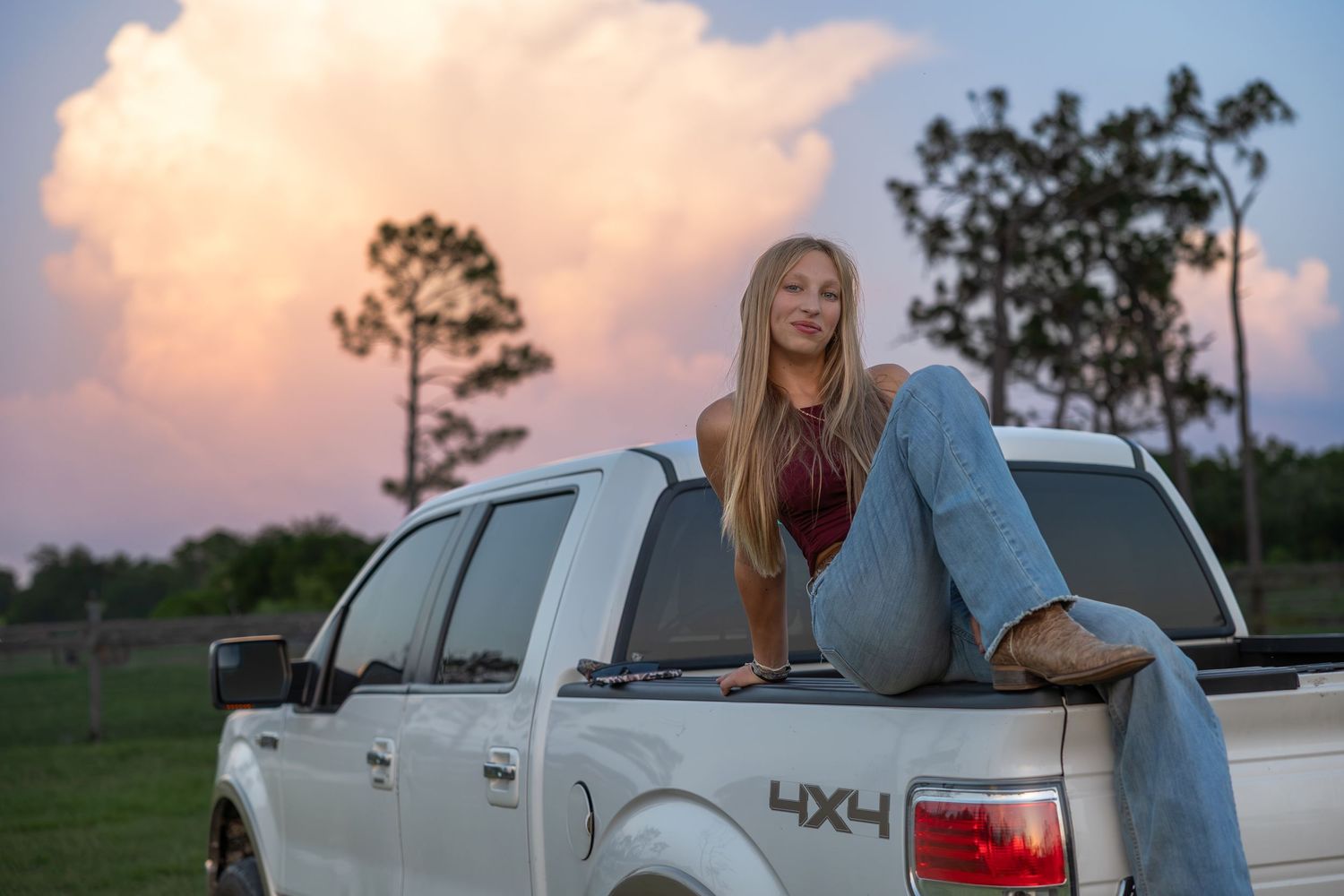 Young woman in jeans sitting on white Ford F-150 truck bed at sunset with dramatic pink clouds. Senior photo session.