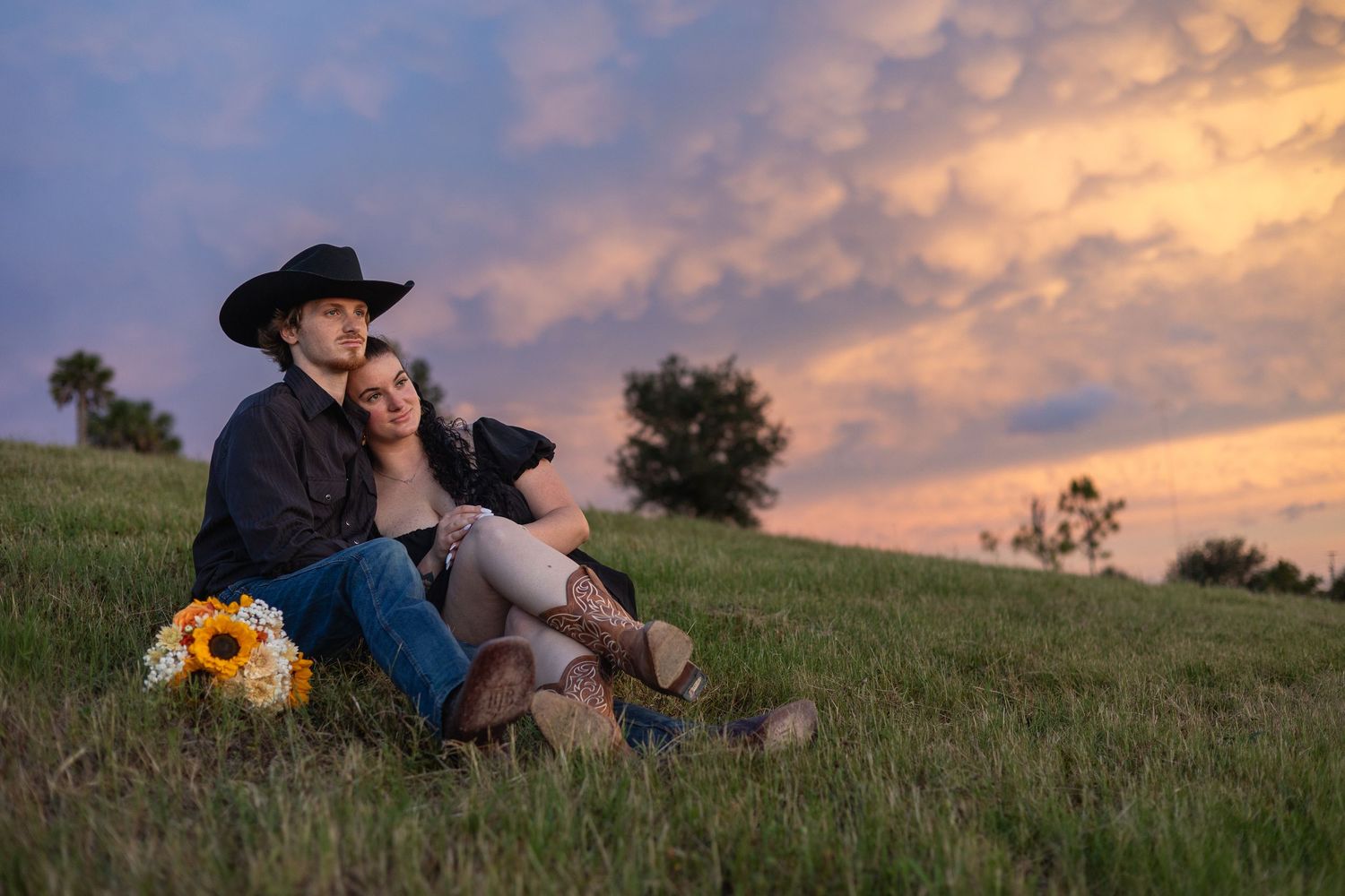 Couple sitting on grassy hill at sunset, man in cowboy hat embracing woman holding sunflowers, romantic countryside engagement photo.