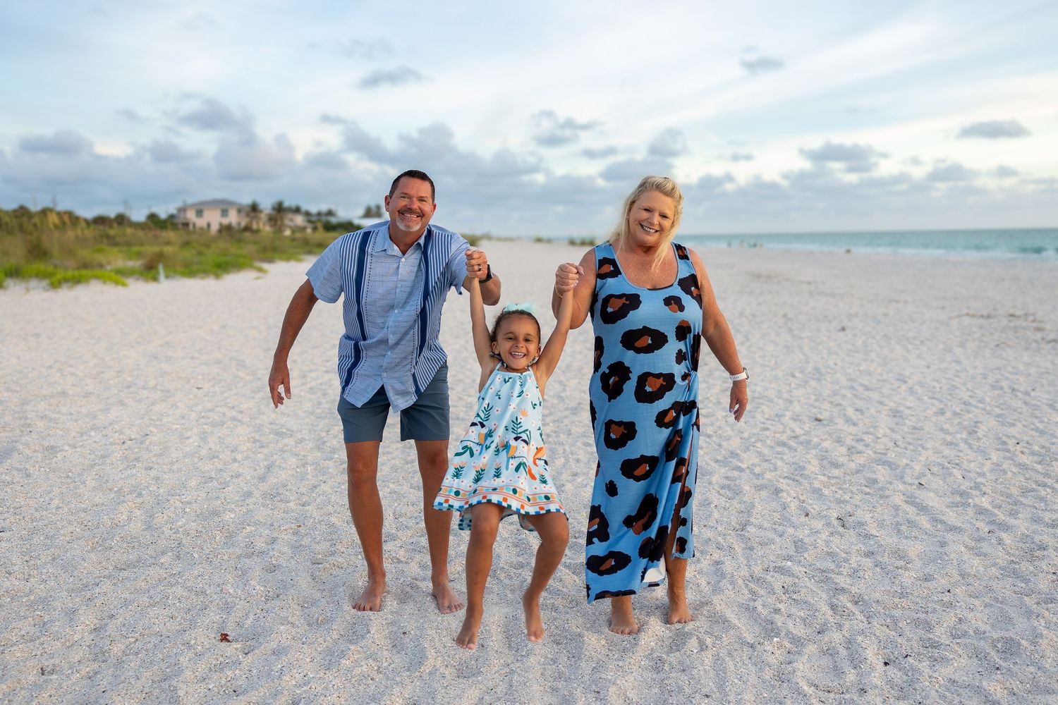 Family of three joyfully swinging child on white sand beach at sunset with turquoise water and cloudy sky.