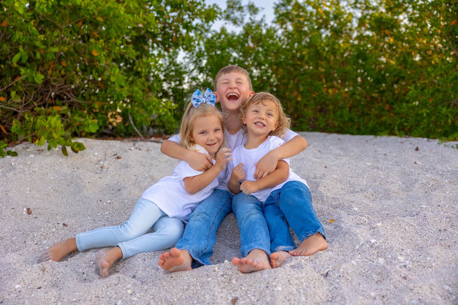Three happy children sitting on sandy beach, wearing white shirts and jeans, laughing and hugging outdoors.