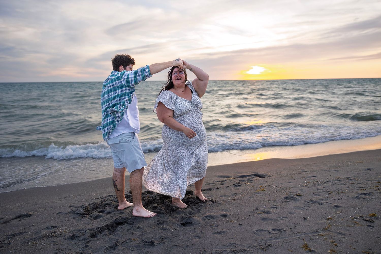 Couple dancing barefoot on a sandy beach at sunset with ocean waves in the background.