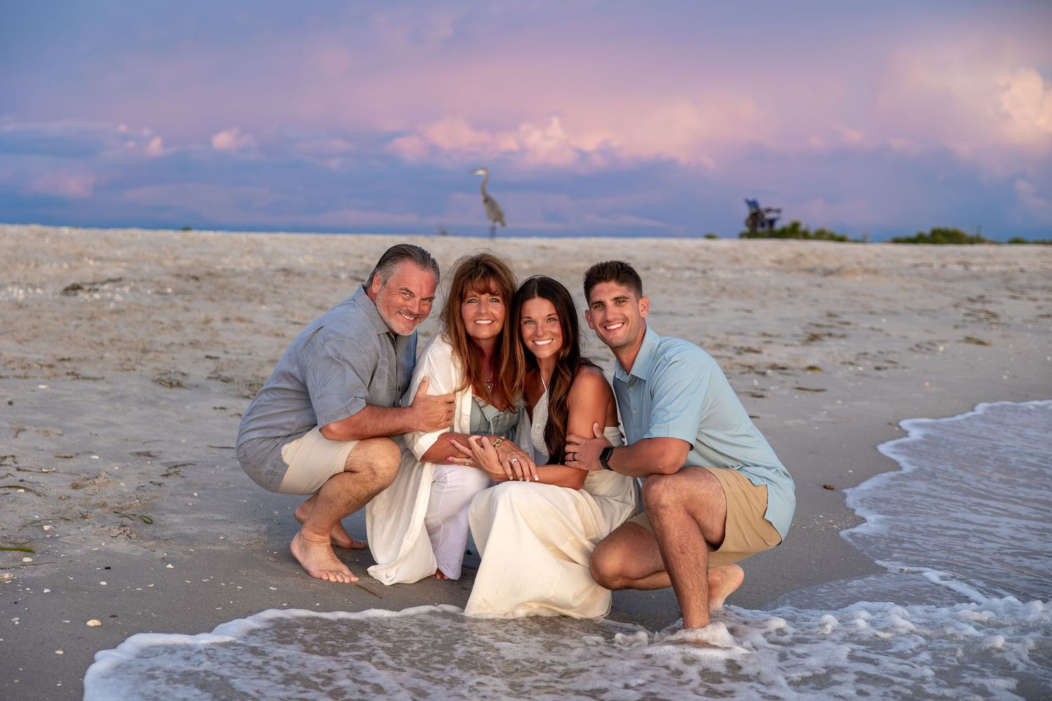 Family of four smiling together on a sandy beach at sunset with colorful pink and purple clouds in the background.