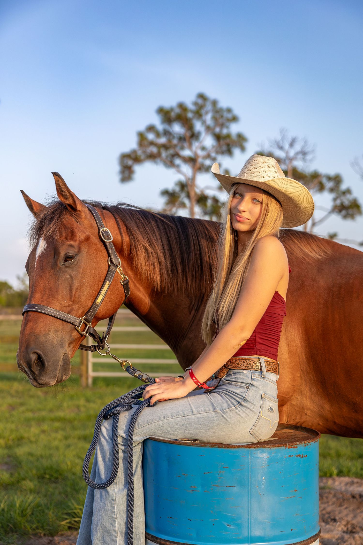 Young blonde woman in cowboy hat and jeans sits on blue barrel beside chestnut horse on sunny farm. Senior photo session.