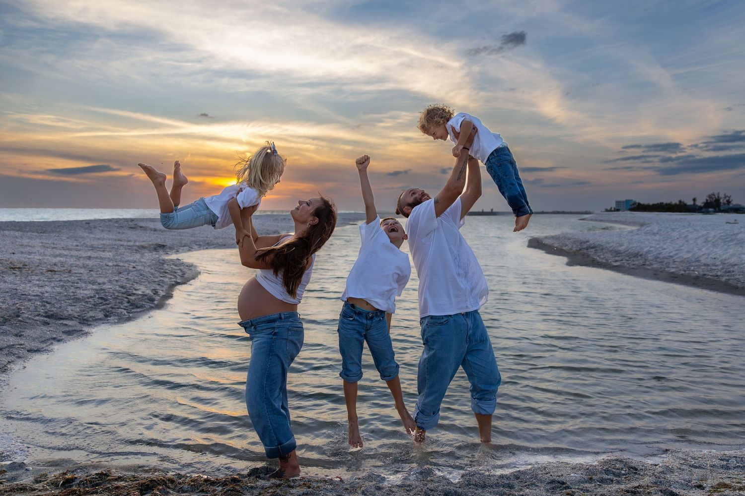 Family of five joyfully playing on a beach at sunset, parents lifting children in the air near ocean waves.