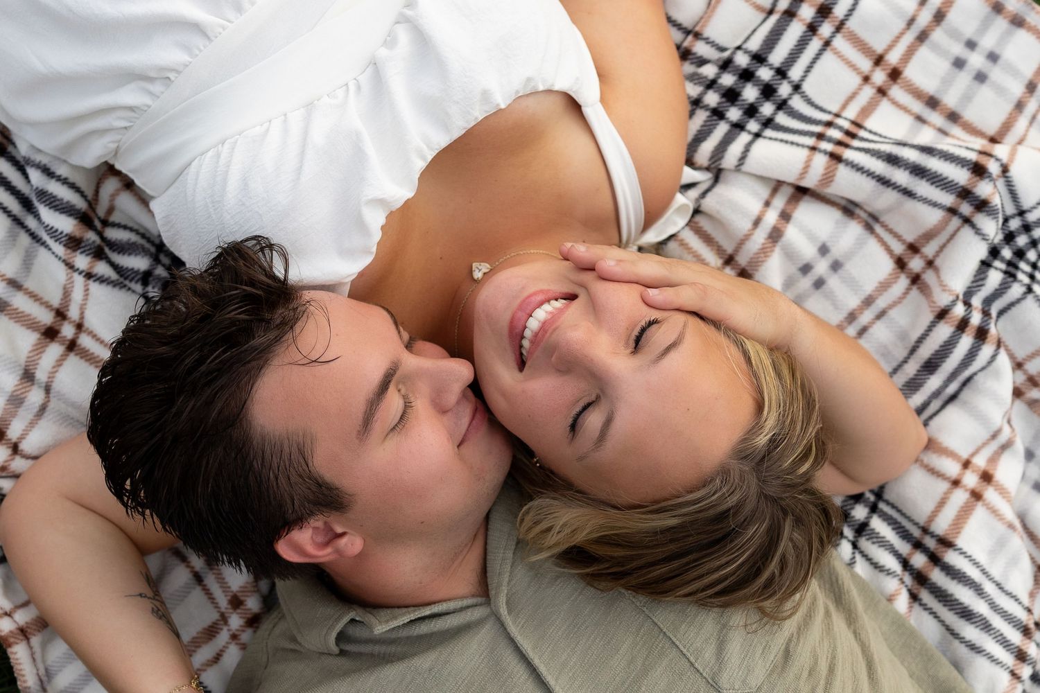 A happy couple lying on a plaid blanket, smiling and gazing at each other in a romantic and intimate moment.