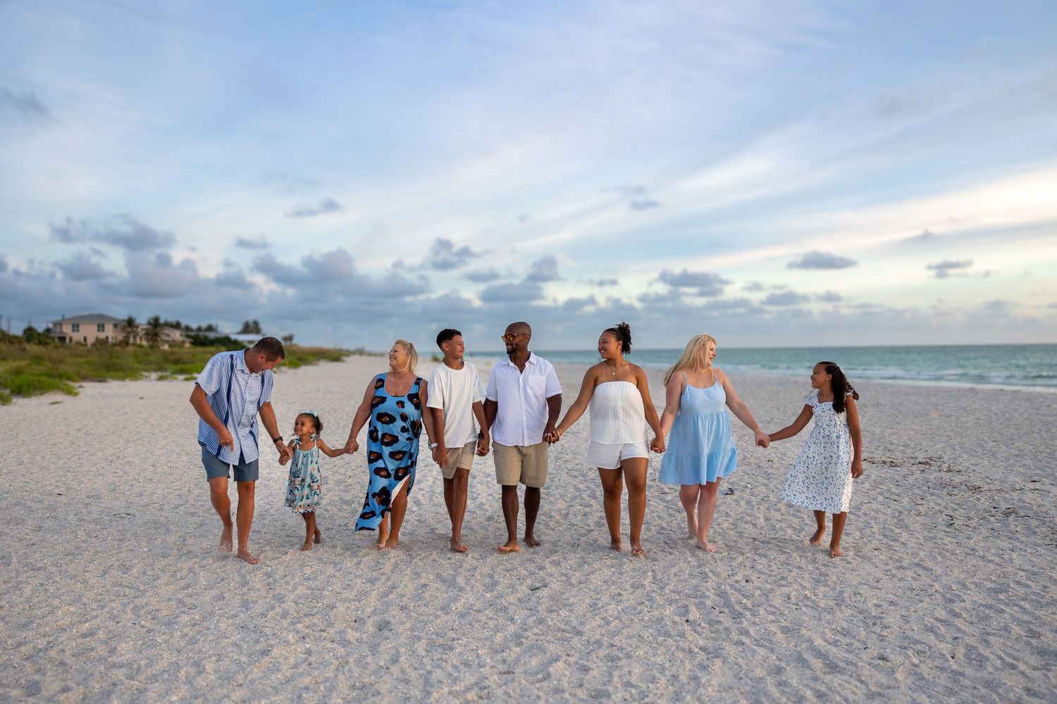 A multigenerational family holds hands walking together on a sandy beach at sunset in matching blue and white outfits.
