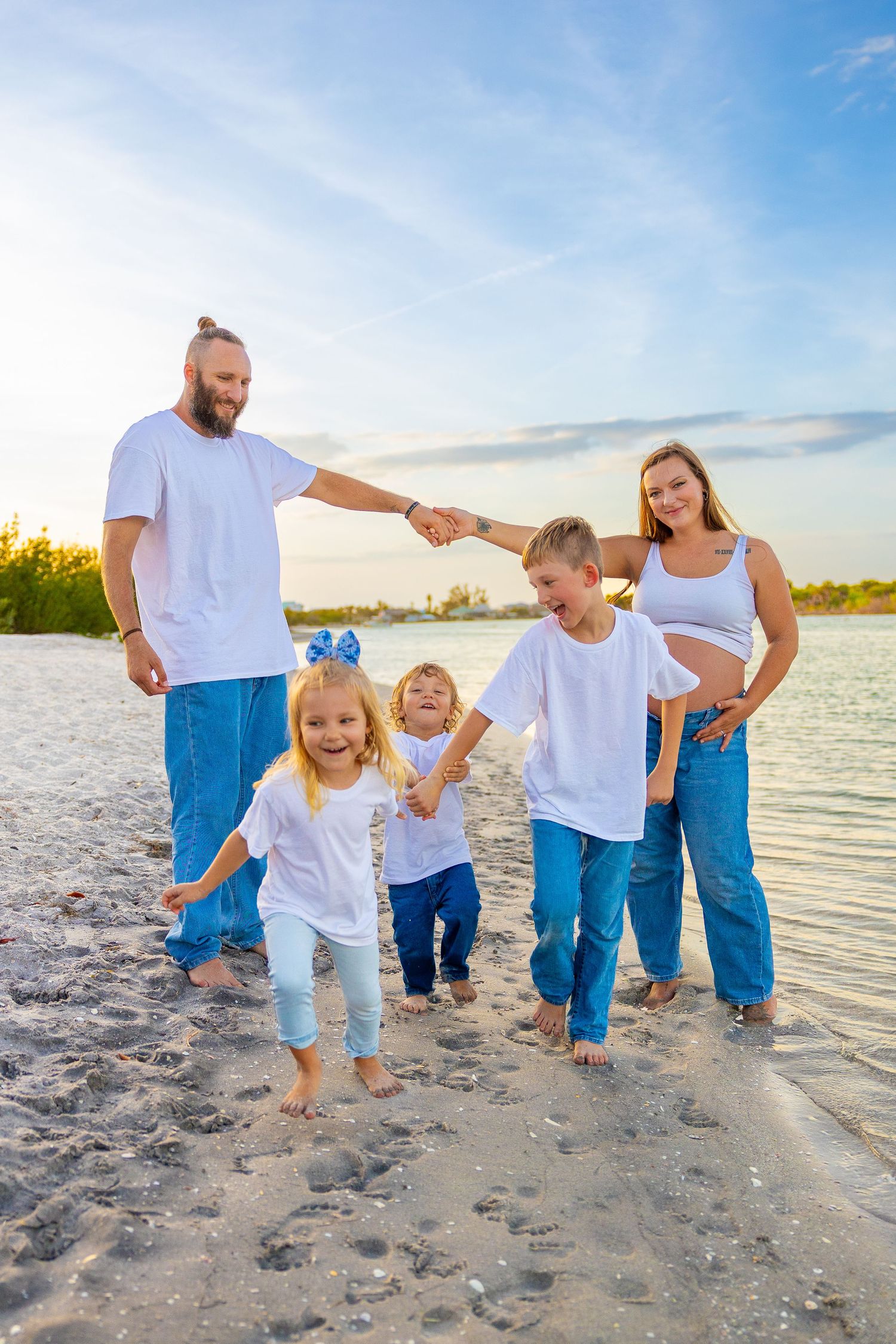 Happy family with three kids holding hands on a sunny beach, wearing matching white shirts and blue jeans.
