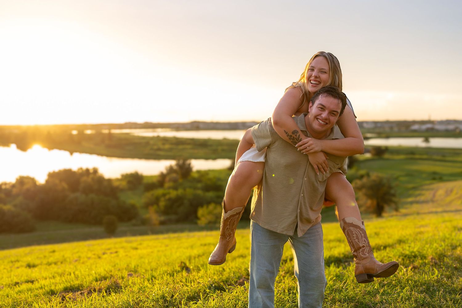 Couple enjoying a piggyback ride on a green hill at sunset with a scenic river and golden sky in the background.
