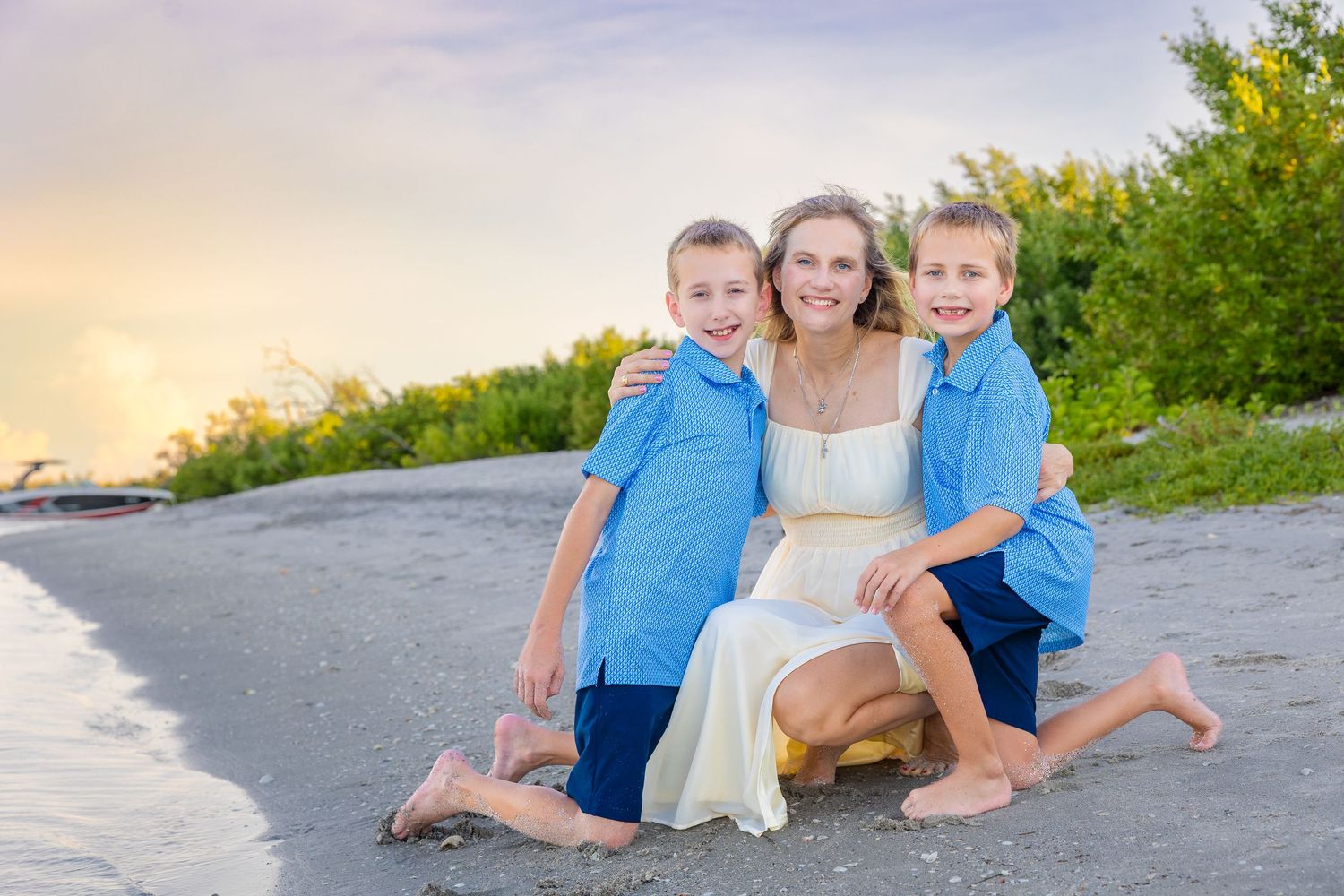 A woman in a white dress smiles on a beach with two boys in blue shirts at sunset.