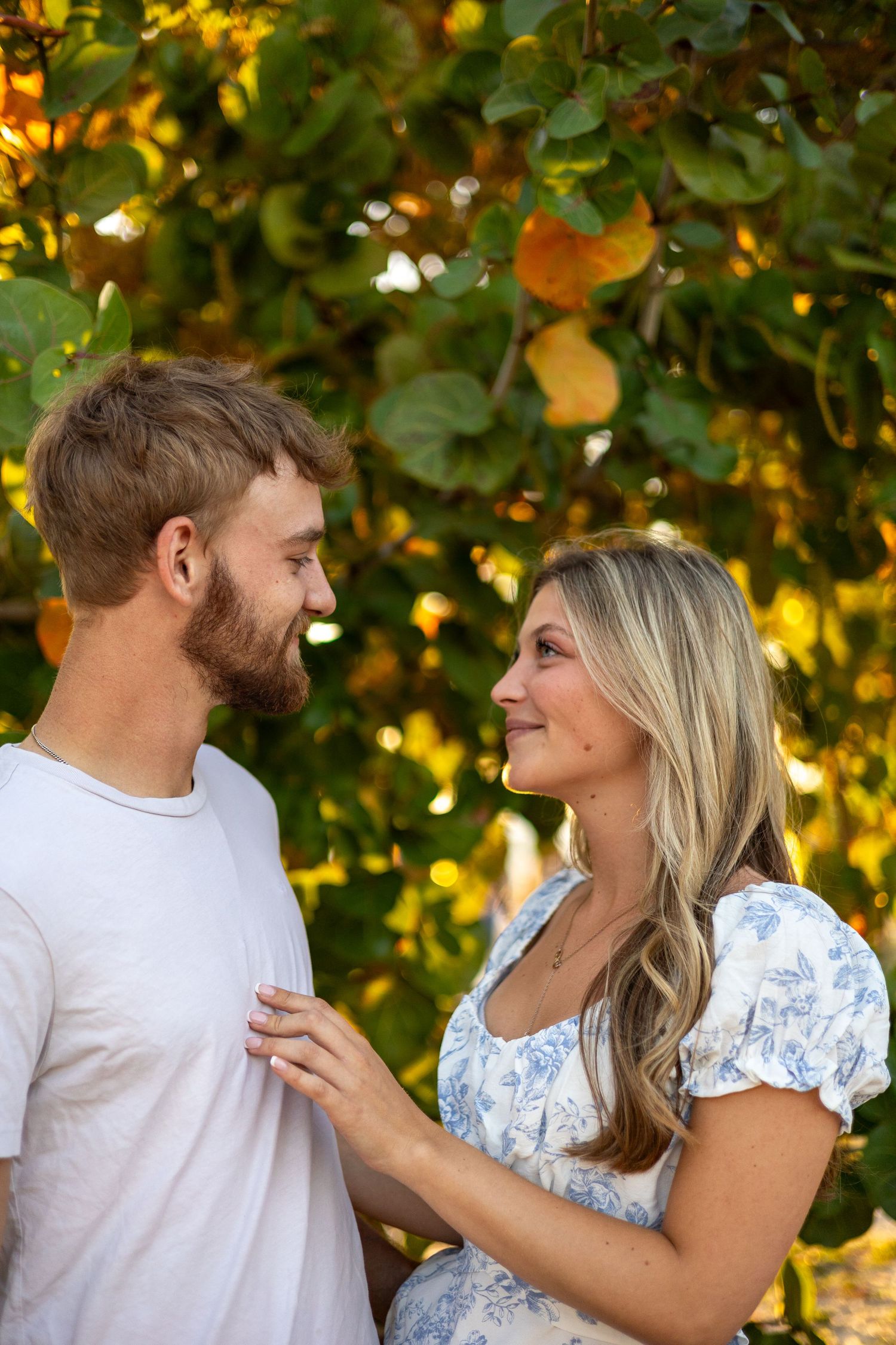 A smiling couple gazes into each other eyes standing before lush green and yellow foliage.