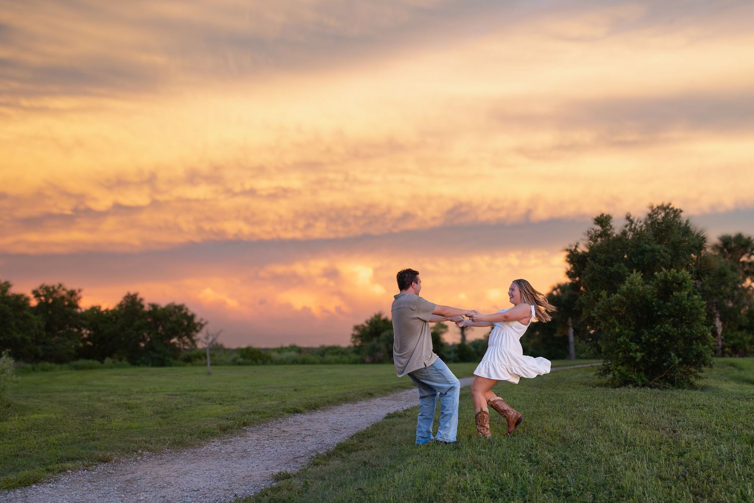 Couple dancing and holding hands on a scenic path at sunset with vibrant orange and pink clouds in the sky.