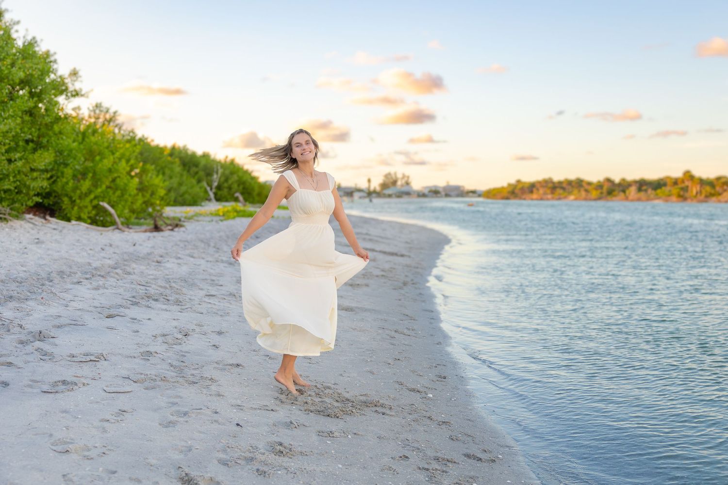A young woman in a flowing white dress walks barefoot along a sandy beach at sunset.