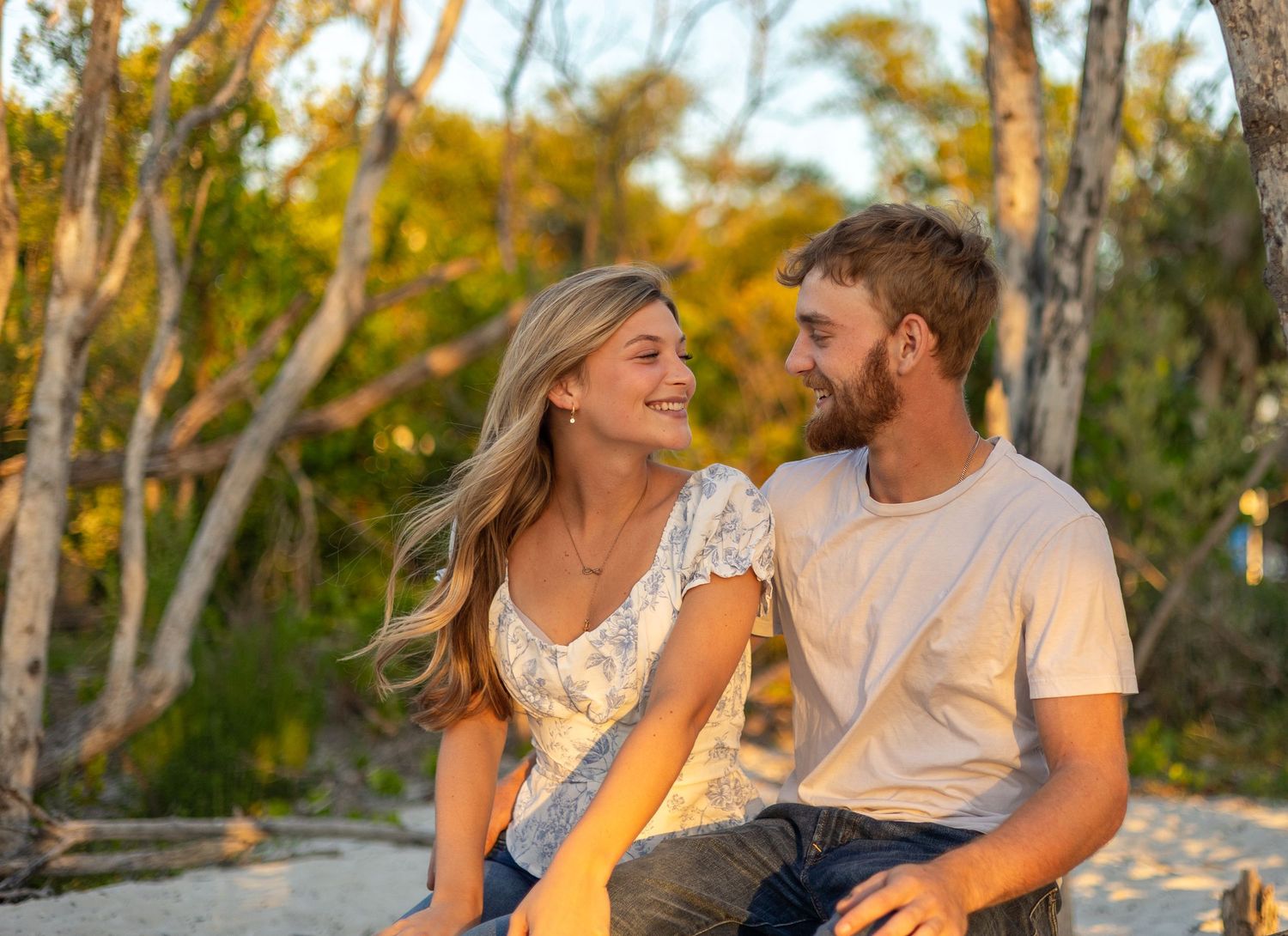 A happy couple sits together on a sandy beach surrounded by trees in warm golden sunset light.