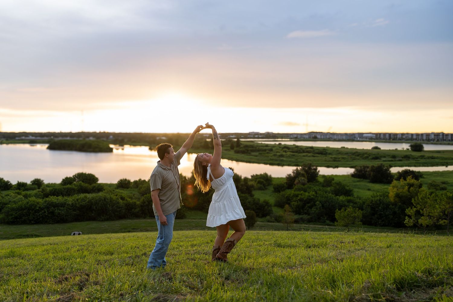 Couple dancing at sunset near a scenic lake surrounded by lush greenery, forming a heart shape with their hands.