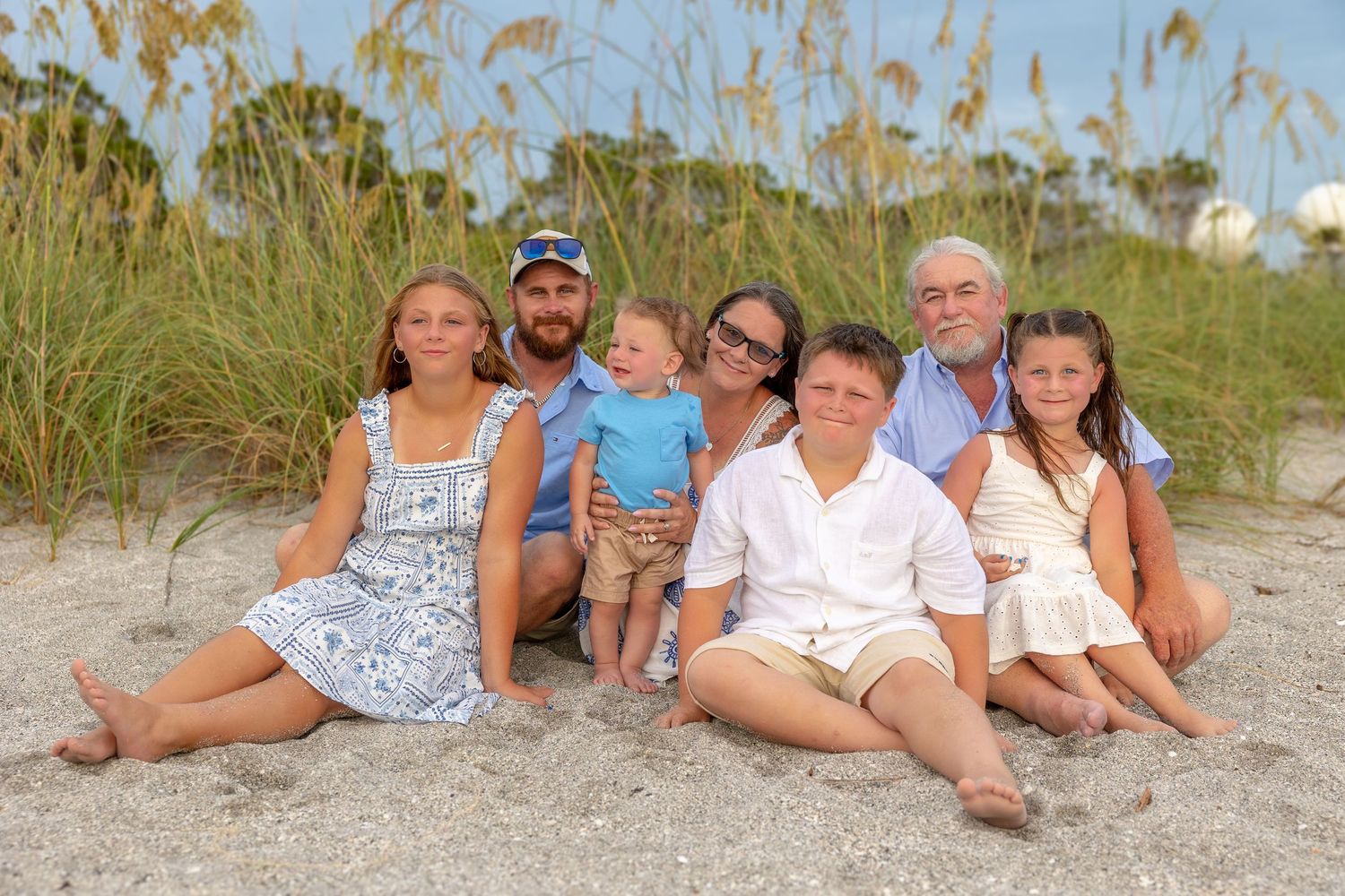 Family of seven posing together on a sandy beach with sea oats in the background on a sunny day.