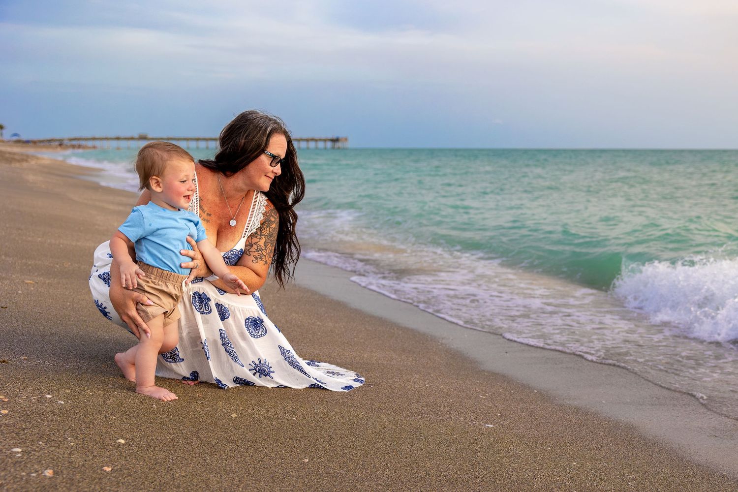 A mother and toddler son enjoy a sunny beach day together, crouching by the ocean waves on a sandy shore.