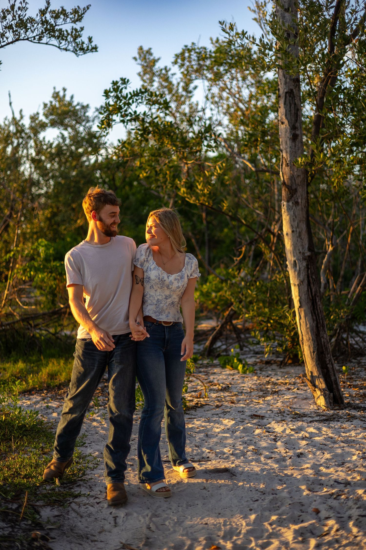 A couple walks hand in hand along a sandy path through green trees in warm golden sunset light.