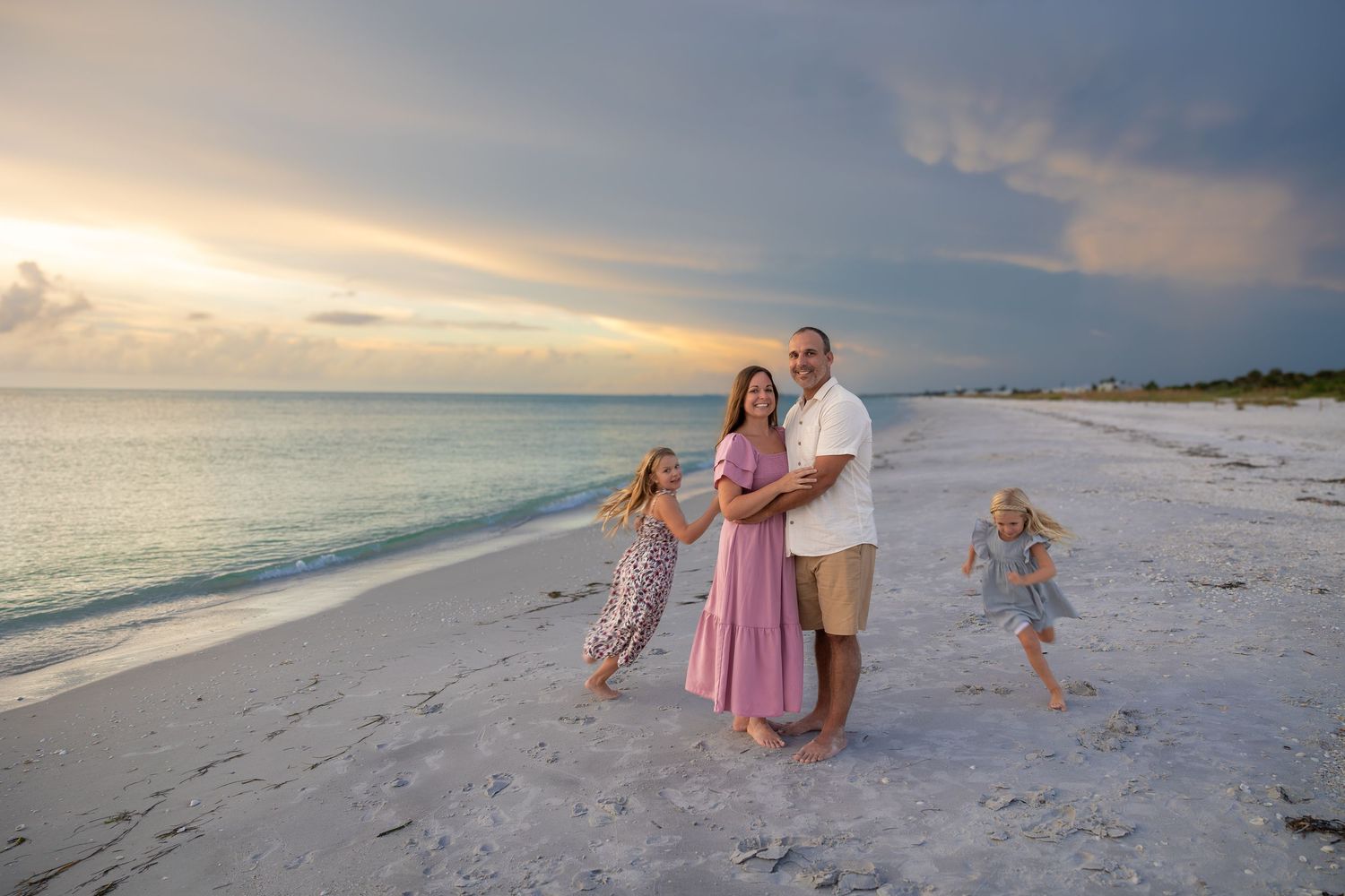 Family posing on a white sand beach at sunset with turquoise water and dramatic colorful sky in background.