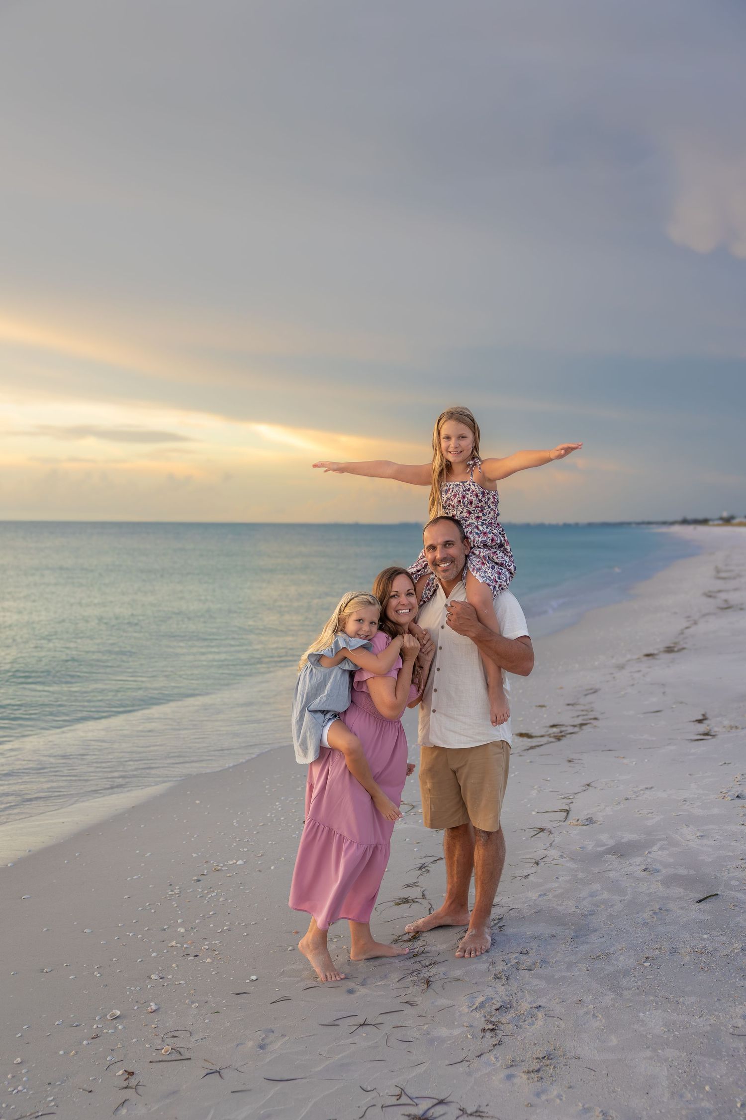 Family of four poses on a sandy beach at sunset, with a girl sitting on dad’s shoulders and arms spread wide.